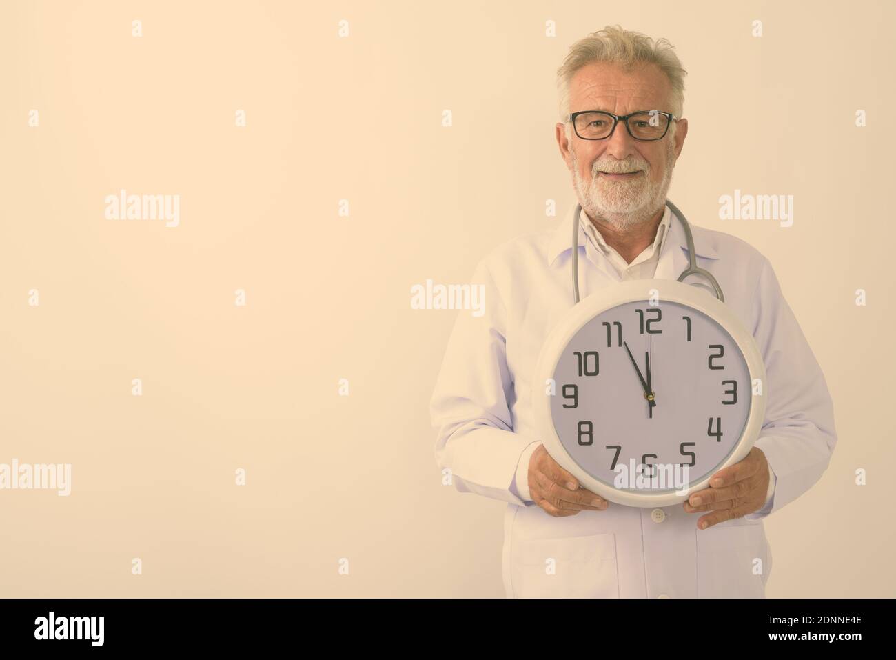 Studio shot of happy senior bearded man doctor smiling while holding wall clock against white background Stock Photo