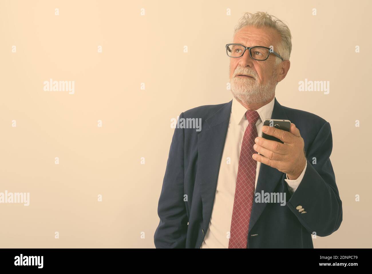 Studio shot of handsome senior bearded businessman thinking while holding mobile phone against white background Stock Photo