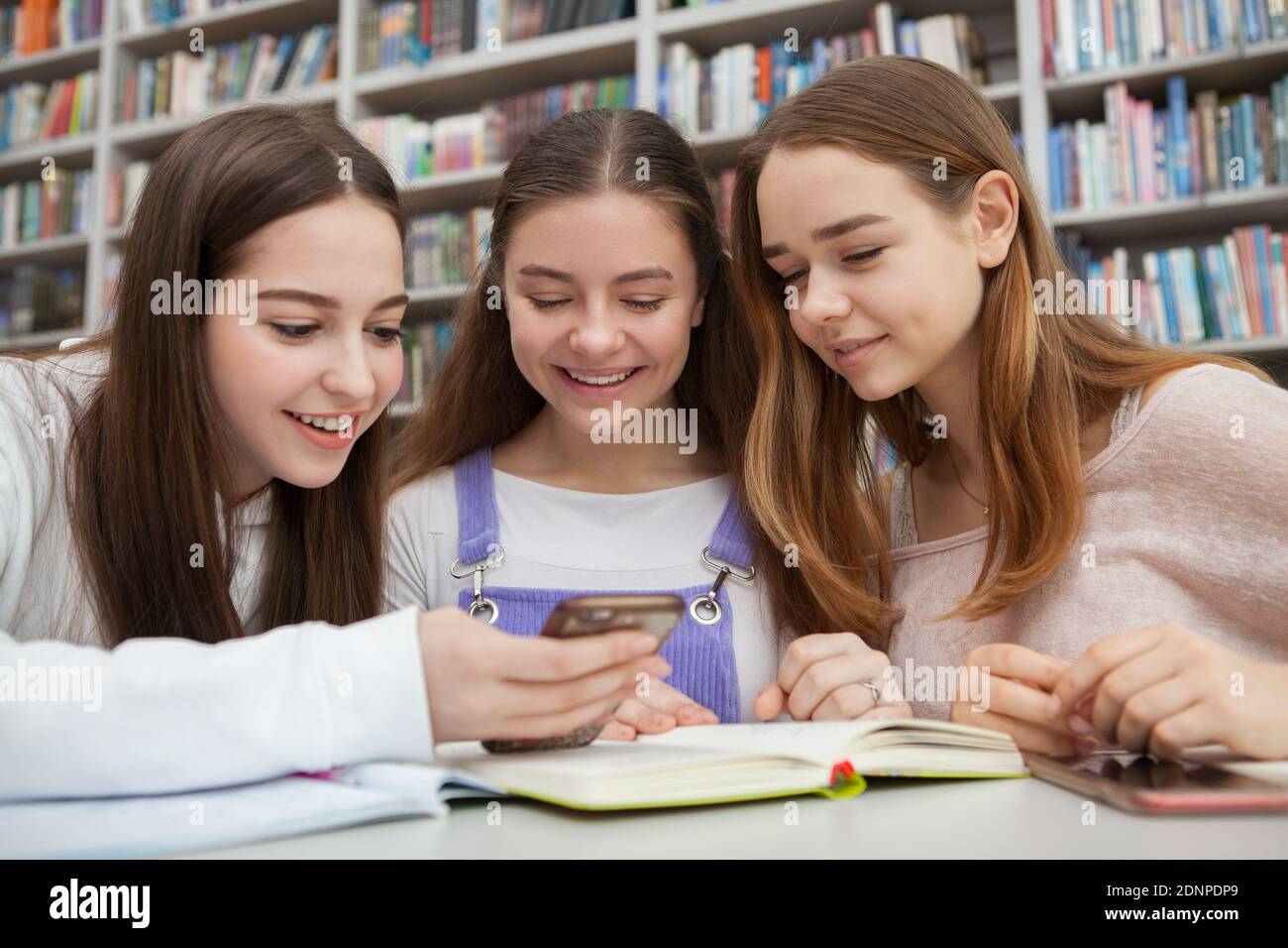 Charming teen girls using smart phone while studying at colege library. Connection, online, social media concept Stock Photo