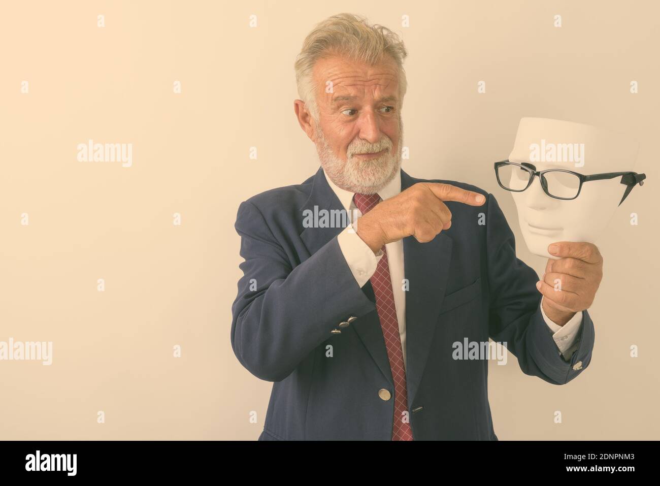 Studio shot of handsome senior bearded businessman looking and pointing at white mask with eyeglasses against white background Stock Photo