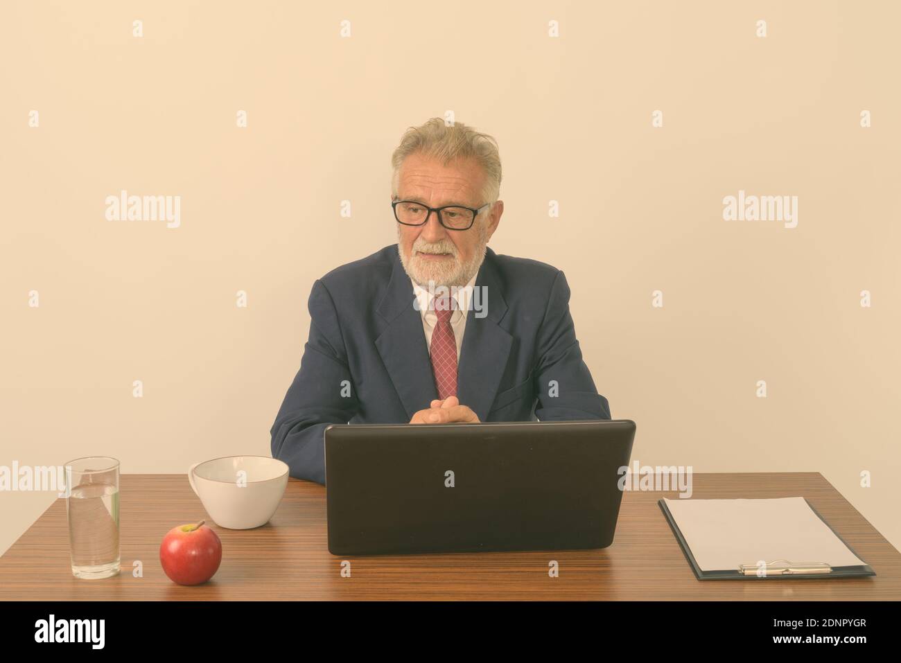 Studio shot of handsome senior bearded businessman thinking while sitting with basic things for work on wooden table against white background Stock Photo