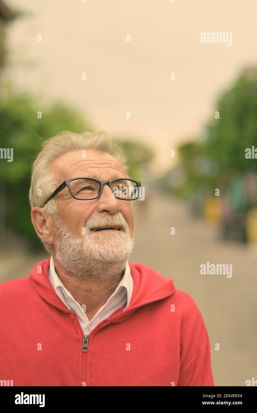 Happy handsome senior bearded man smiling while thinking and looking up with eyeglasses outdoors Stock Photo