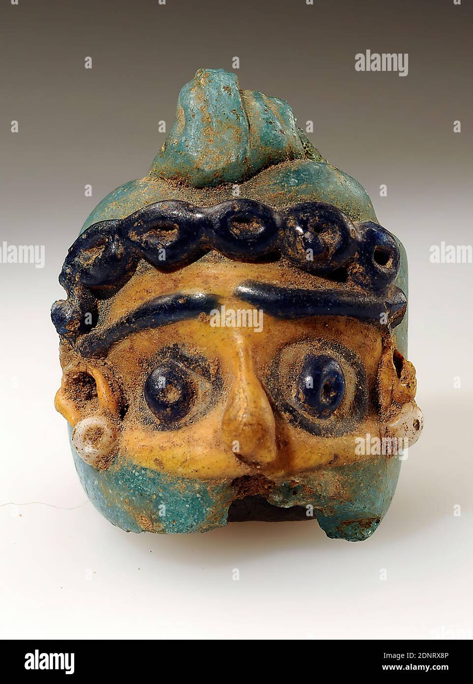 mask bead, glass, mouth-blown, glass paste, Total: Height: 2.4 cm; Width: 2 cm; Depth: 1.9 cm, Small sculptures/small-scale art, Man, non-Christian religions and cults (in general), Antiquity, The bead consists of a turquoise cylinder open at the bottom, horizontally set off at the top and provided with an eyelet grooved in the longitudinal direction, on which a face is placed at the front made of yellow, dark blue and white glass mass. The face itself is yellow, the forehead ends a row of five dark blue ringlets. Stock Photo