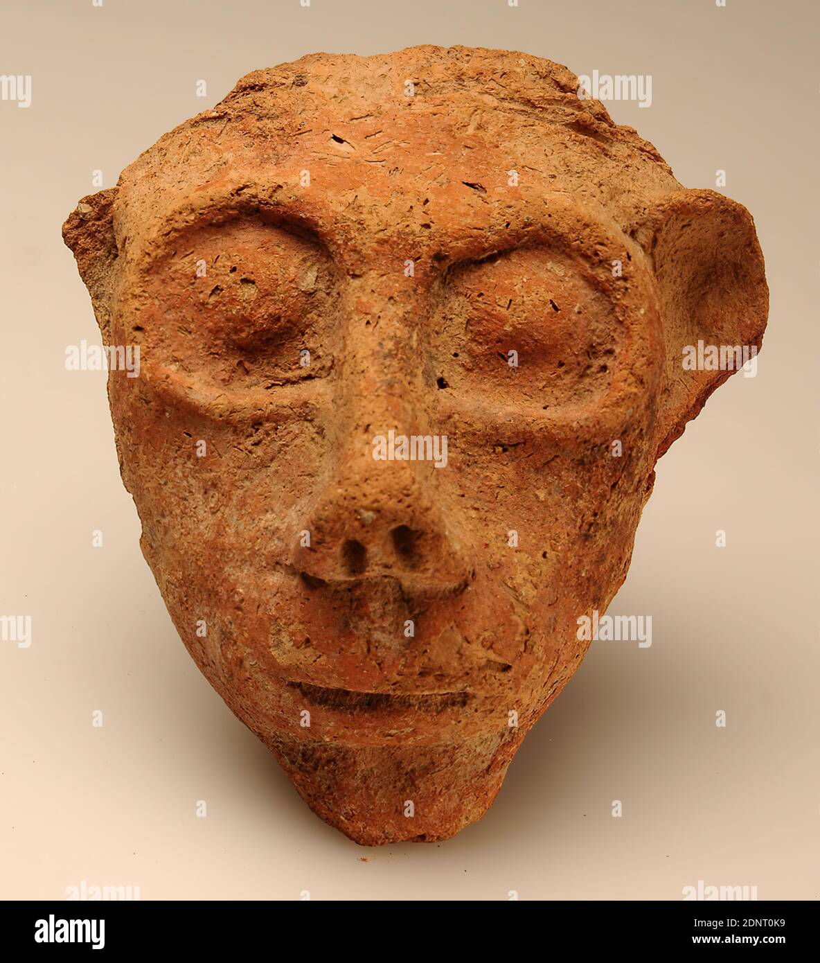 Small head, clay, hand modelled, clay, hand modelled, Total: Height: 7.7 cm; Width: 7.4 cm; Depth: 4.2 cm, ceramics, small sculptures, antiquity, The frontal view of the head is characterized by large, almost round eye sockets with a circumferential arch, the protruding, widely projecting left ear and a narrow, horizontally incised mouth. The nose is a little pre-bent. The cheeks are flat and don't step out. Below the chin, the base of the neck is preserved. Above the forehead there are two horizontal incised lines each indicating hair or headgear. A hole on the back served for fastening. Stock Photo