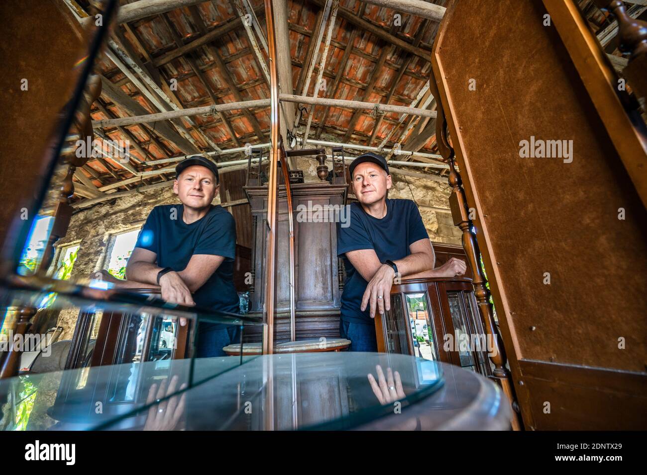 Reflection in the mirror of a man looking into the camera in a storage room full of old wooden furniture Stock Photo