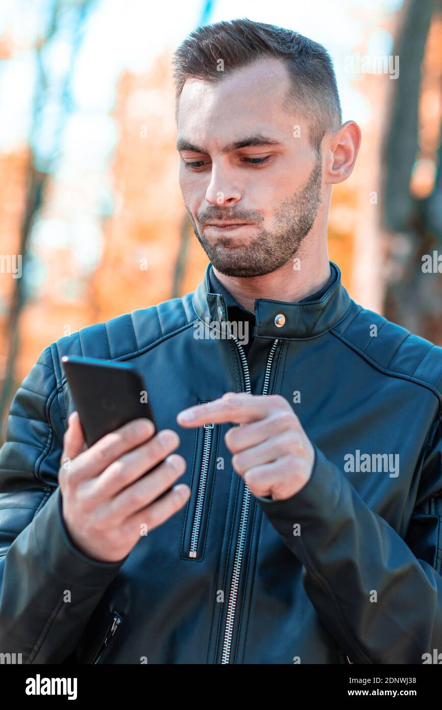 Youthful Guy Using Black Smartphone at the Beautiful Autumn Park. Handsome Young Man with Mobile Phone at Sunny Day - Medium CloseUp Shot Stock Photo