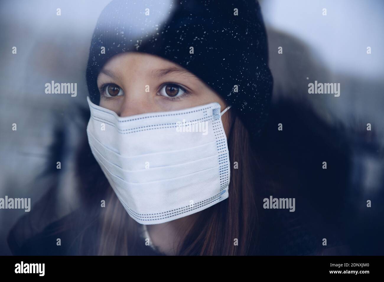 A pretty girl is standing by the window in a protective mask. Stock Photo