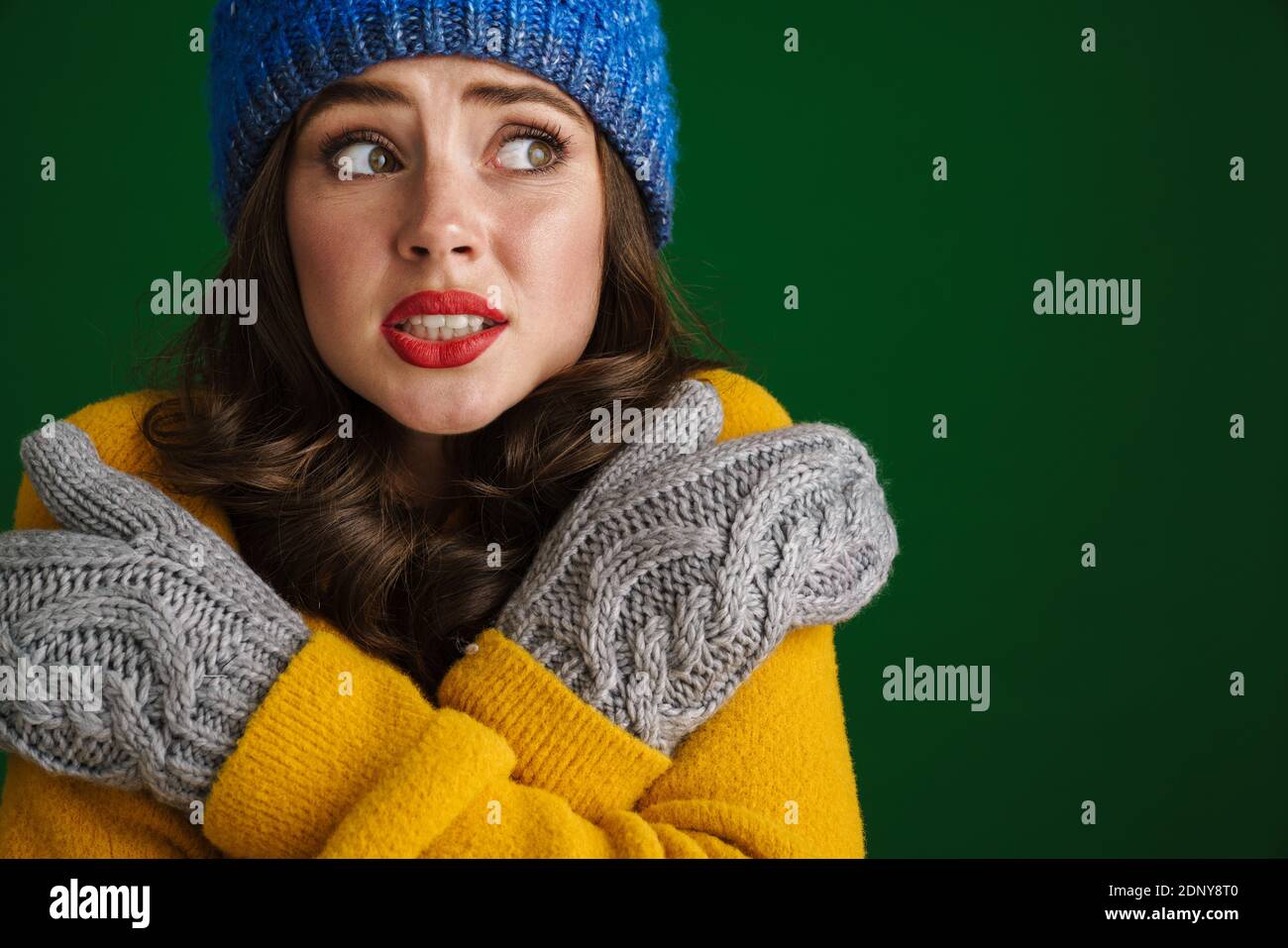 Beautiful unhappy girl in knit hat and mittens posing and trembling isolated over green background Stock Photo