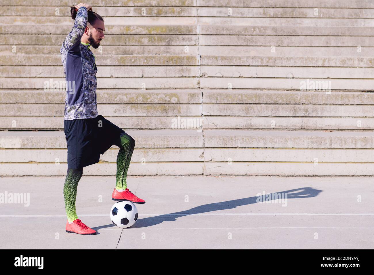 sportsman picking up his hair in a tail with a foot on his football ball in a concrete soccer court, concept of healthy lifestyle and urban sport in t Stock Photo