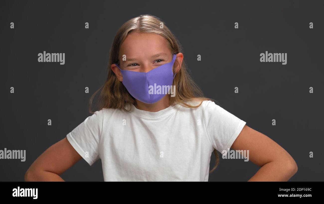 School girl wearing blue reusable face mask wearing white t-shirt smiling with her eyes looking at camera while standing in studio. Isolated on black Stock Photo