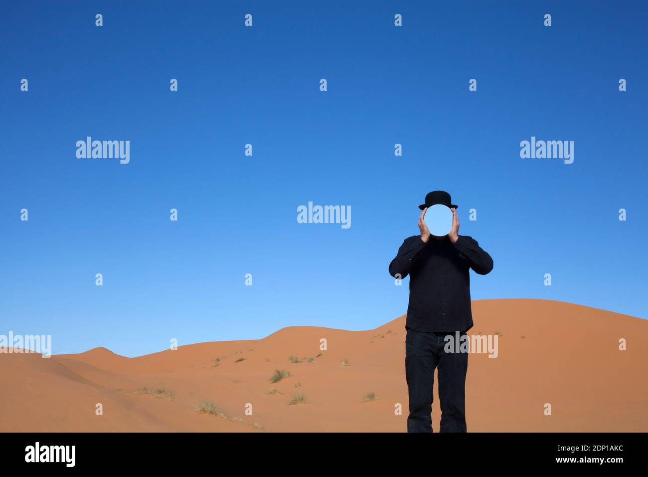 Morocco, Merzouga, Erg Chebbi, man wearing a bowler hat holding mirror in front of his face in desert dune Stock Photo
