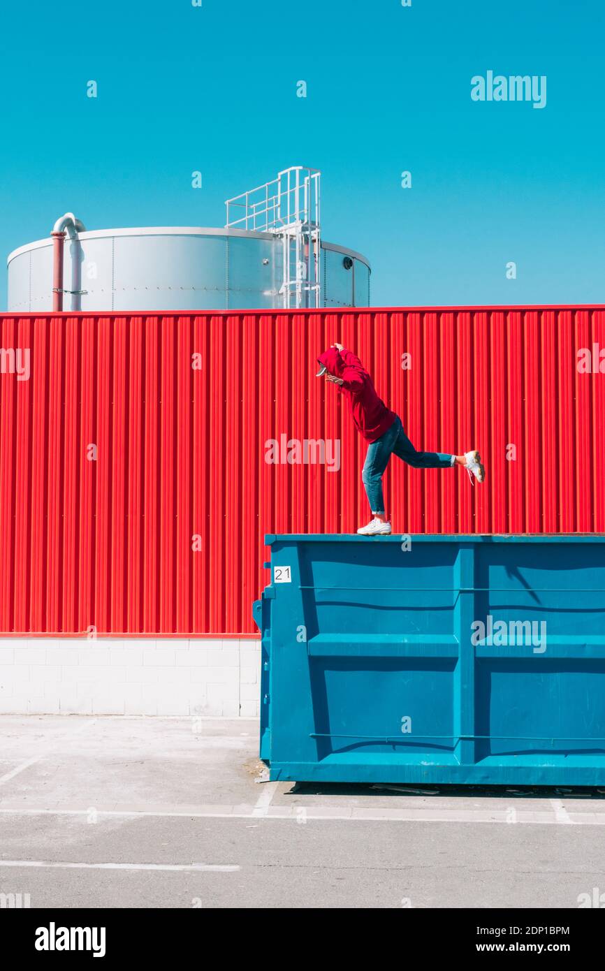 Young man wearing red hooded jacket balancing on edge of container in front of red wall in industrial setting Stock Photo