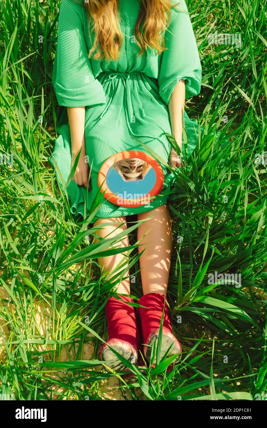 Crop view of young woman wearing green dress sitting on a field looking at mirror Stock Photo
