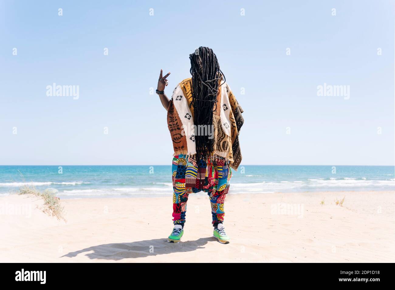 Man standing on beach with dreadlocks hiding his face, making rude hand sign Stock Photo