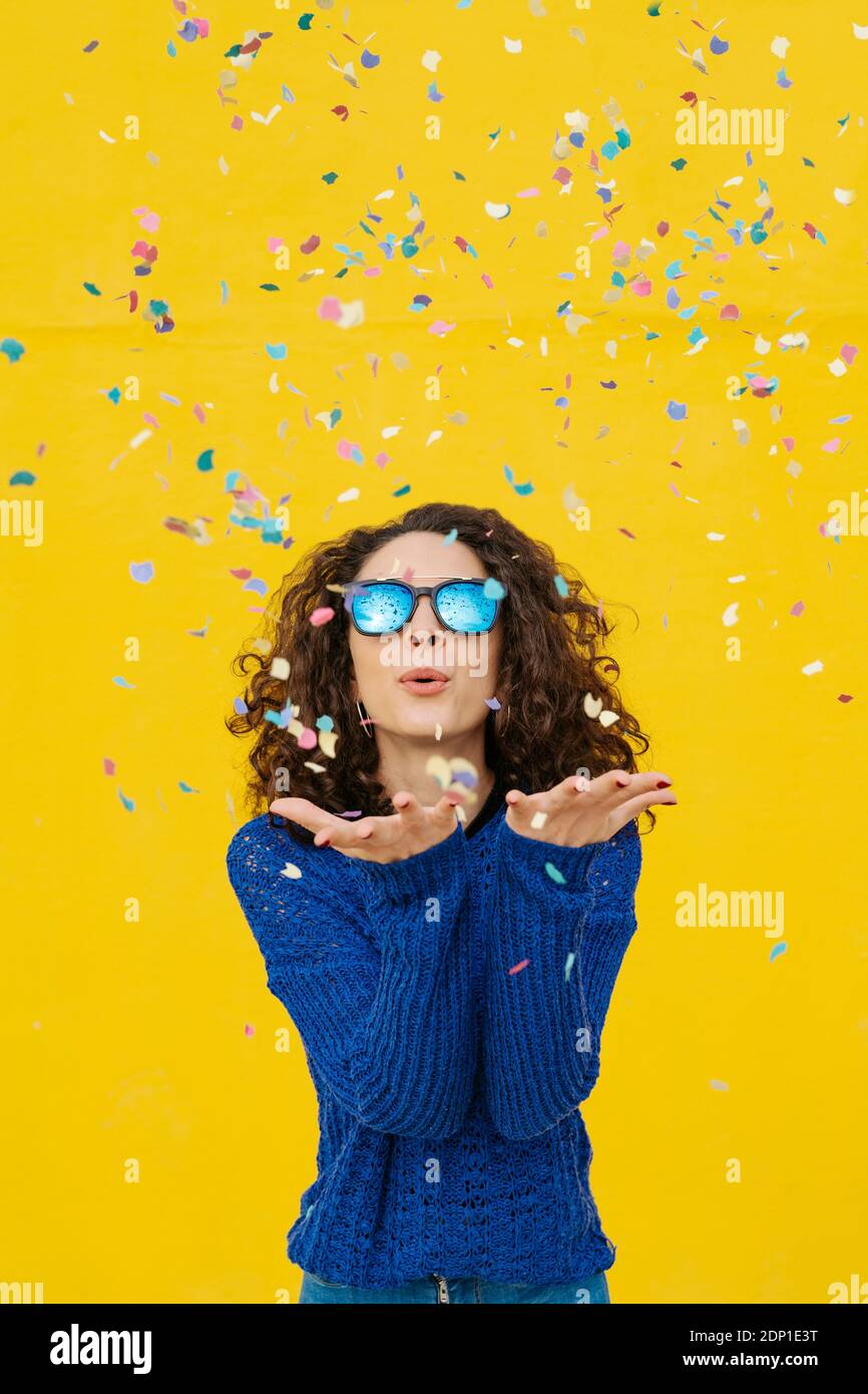 Portrait of young woman with mirrored sunglasses blowing confetti in the air in front of yellow background Stock Photo
