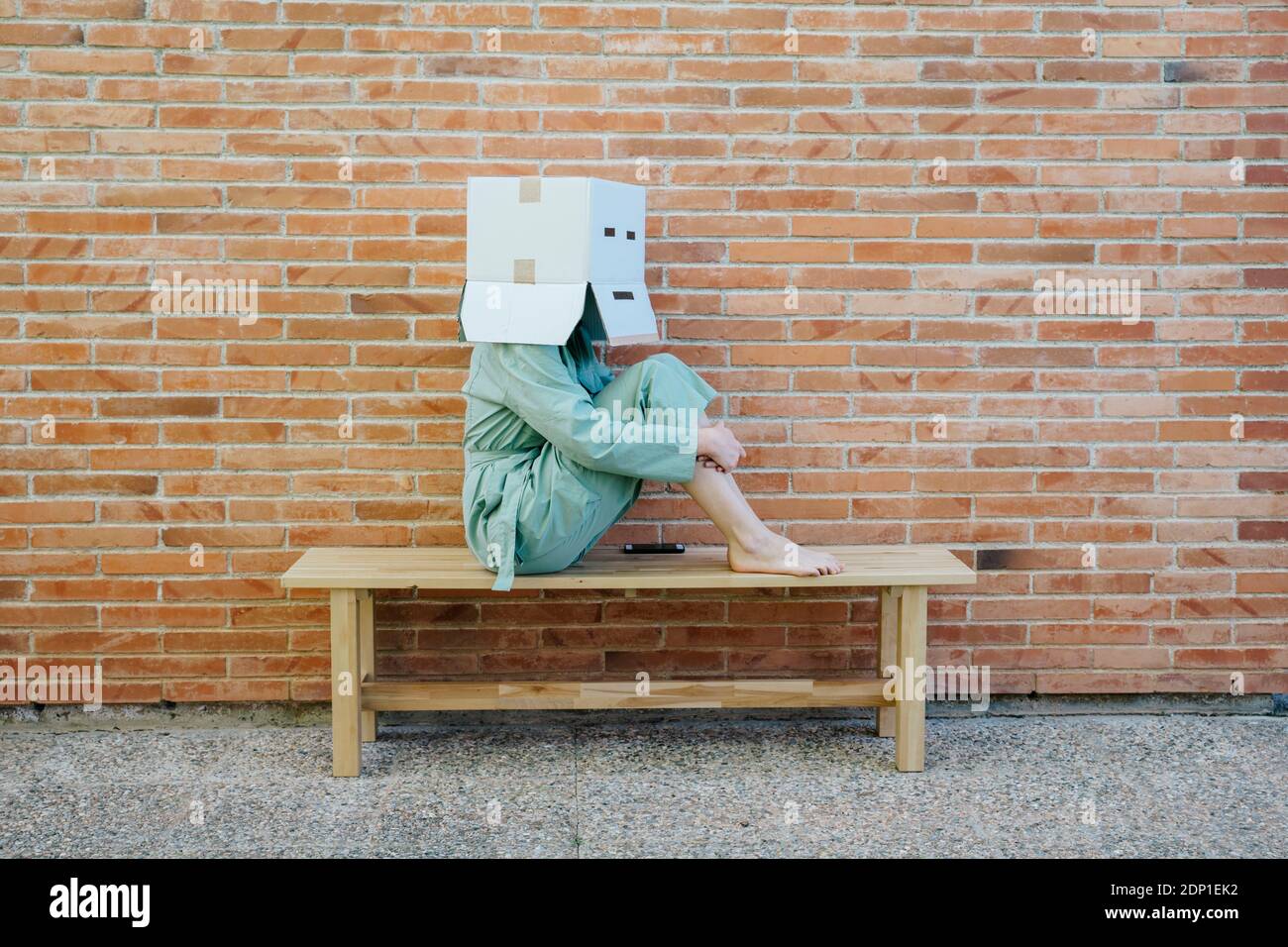 Woman sitting on bench in front of brick wall with serious  face on cardboard box, hugging knees Stock Photo