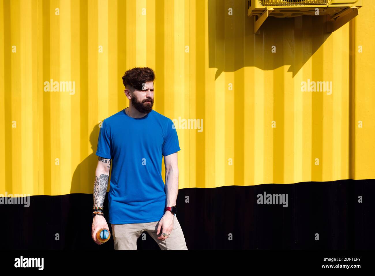 Young man with tattoo standing in front of yellow wall, holding water bottle Stock Photo