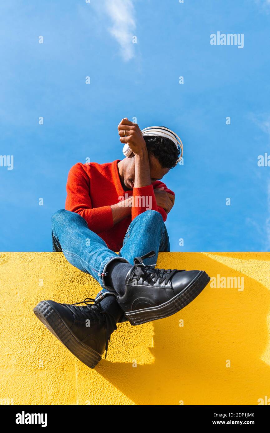 Young man with headphones, sitting on yellow wall, listening music Stock Photo