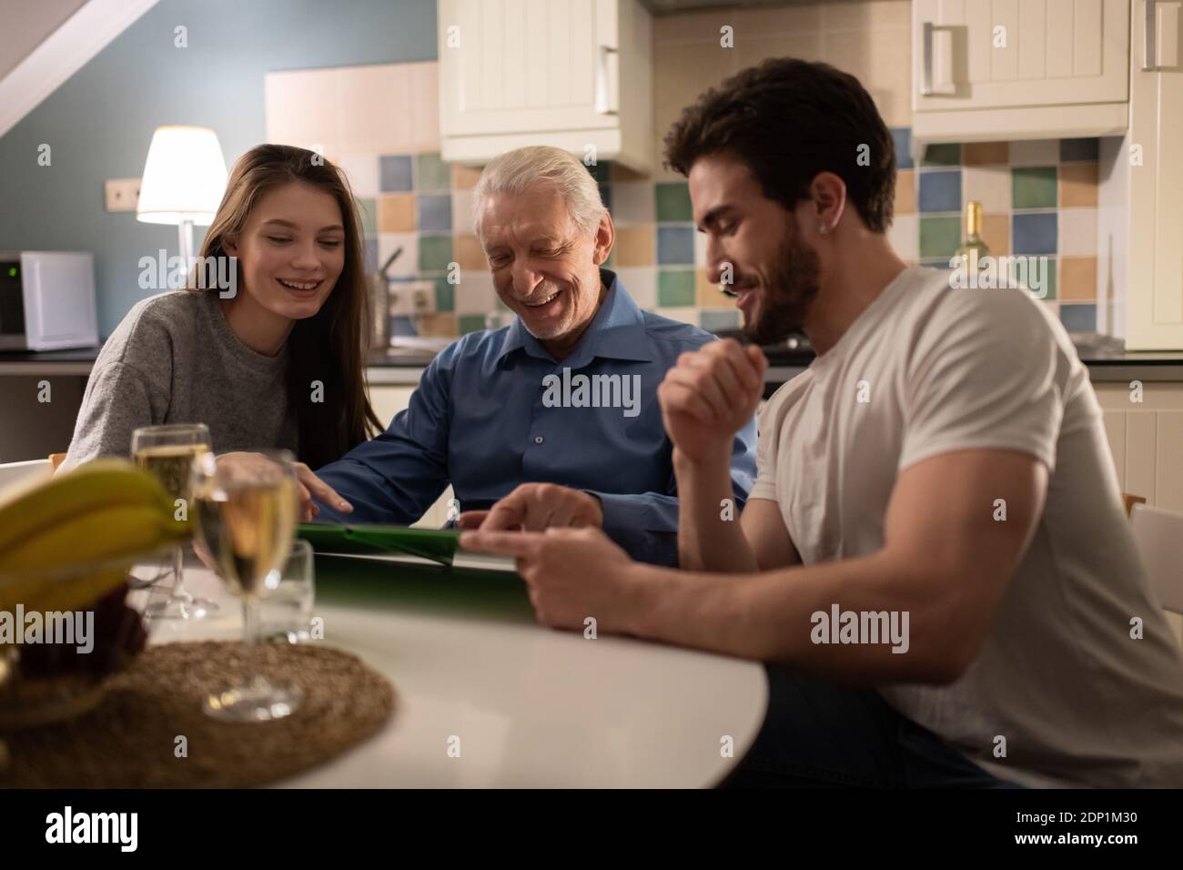 Cheerful mature male smiling and demonstrating family photos to son and daughter in law while sitting in kitchen during dinner Stock Photo