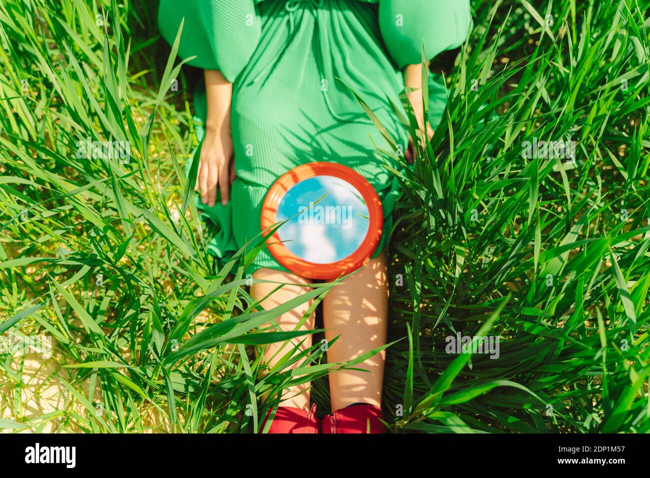 Crop view of young woman wearing green dress sitting on a field with mirror reflecting clouds Stock Photo