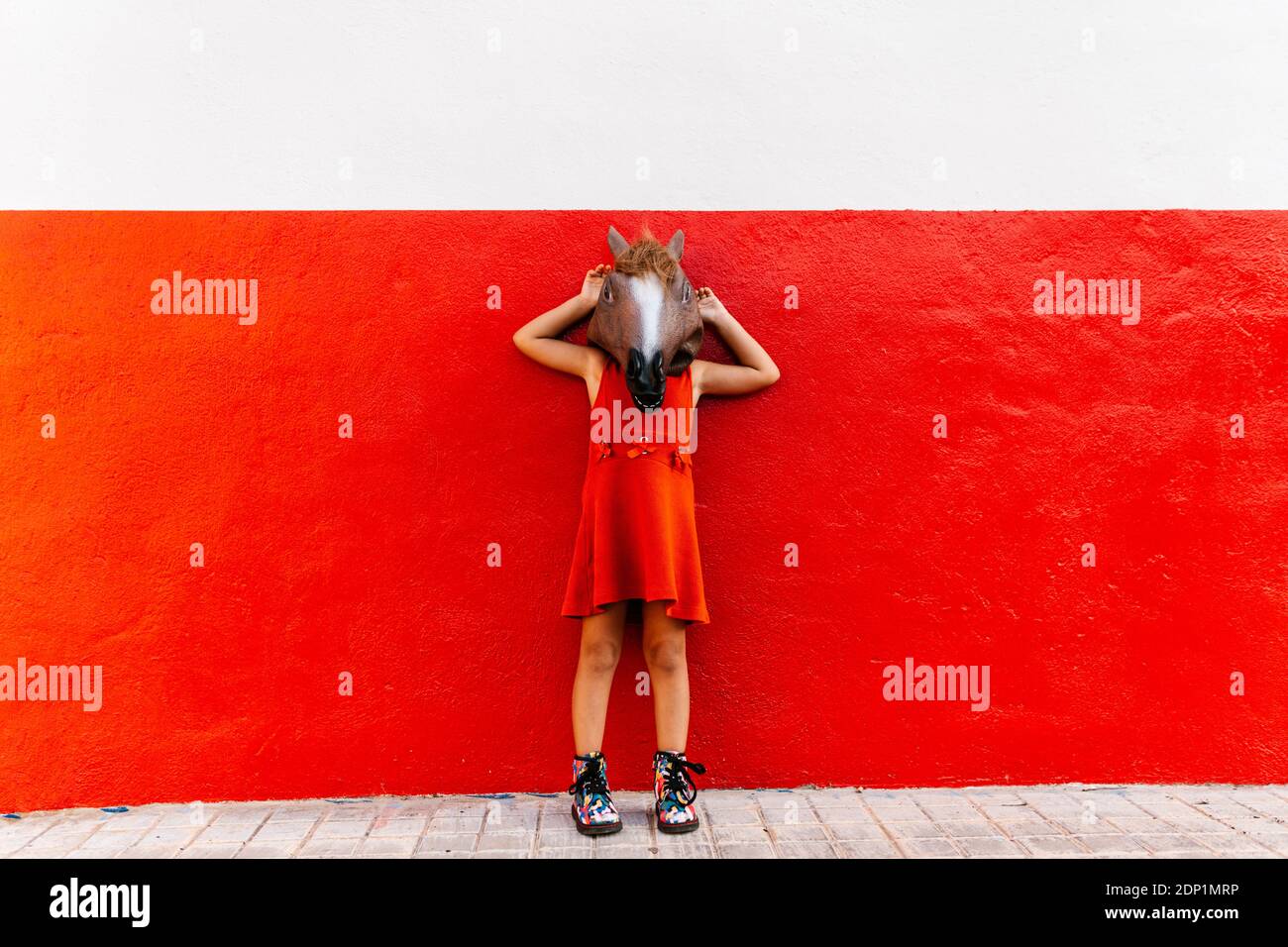 Little girl with a horse's head and a red dress Lening on a red and white wall Stock Photo