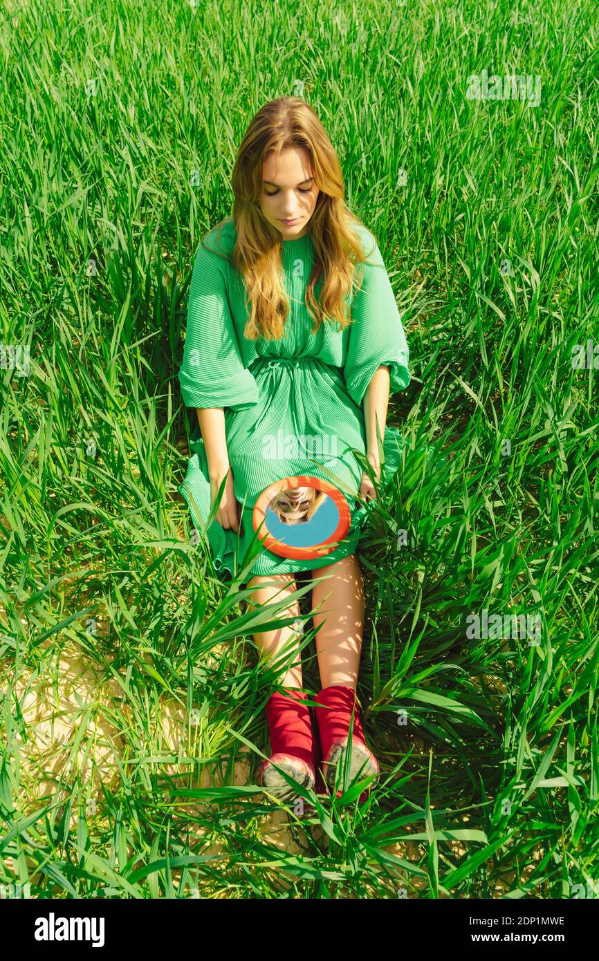 Young woman wearing green dress sitting on a field looking at mirror Stock Photo