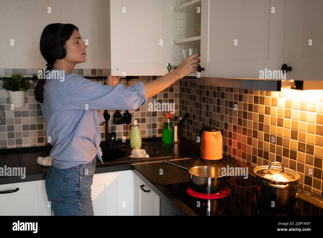 Side view of casual female in headphones putting clean glass in cupboard while cleaning kitchen at home and listening to music Stock Photo