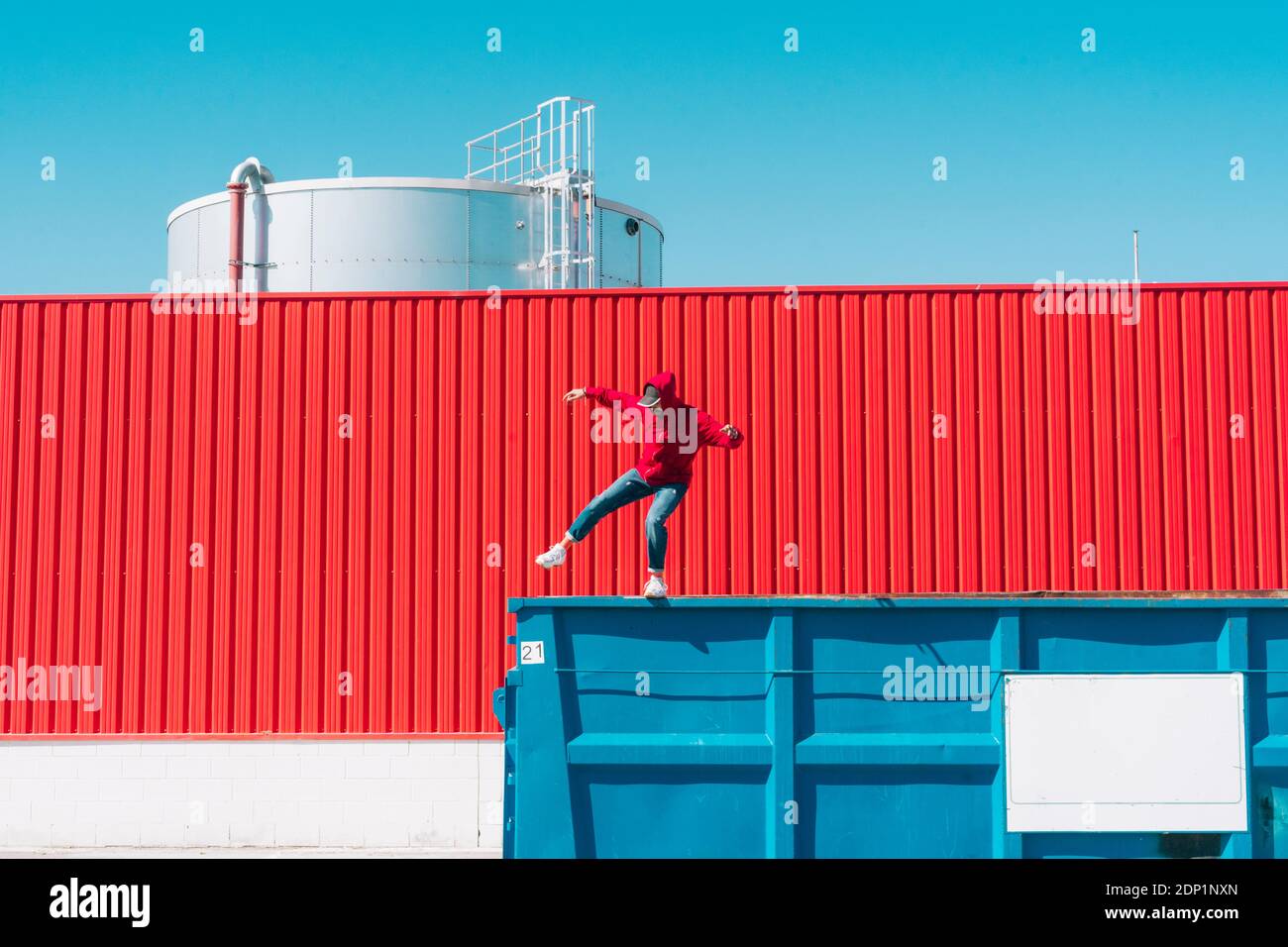 Young man wearing red hooded jacket balancing on edge of container in front of red wall in industrial setting Stock Photo