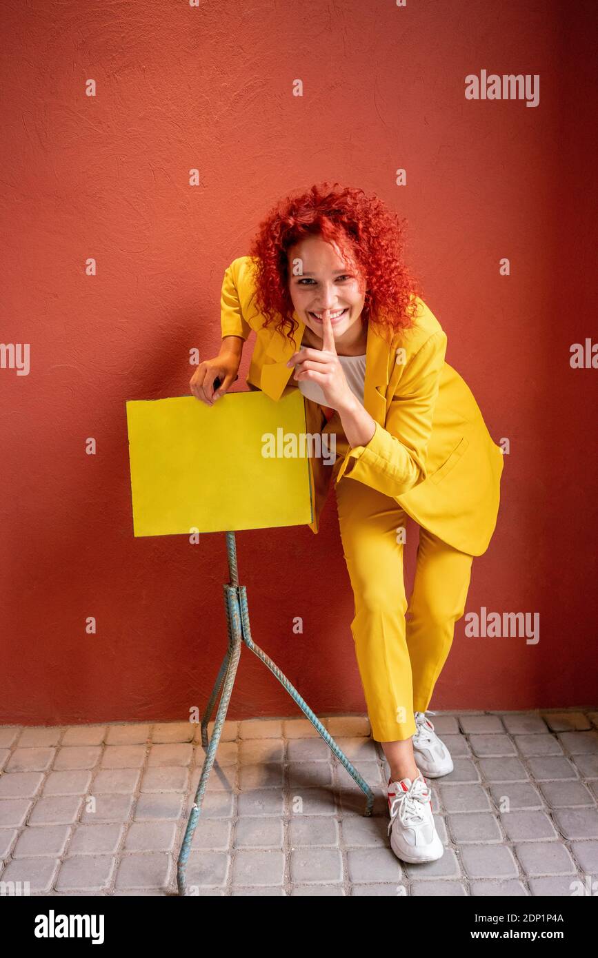 Young woman  in yellow suit leaning on music stand with finger on mouth Stock Photo