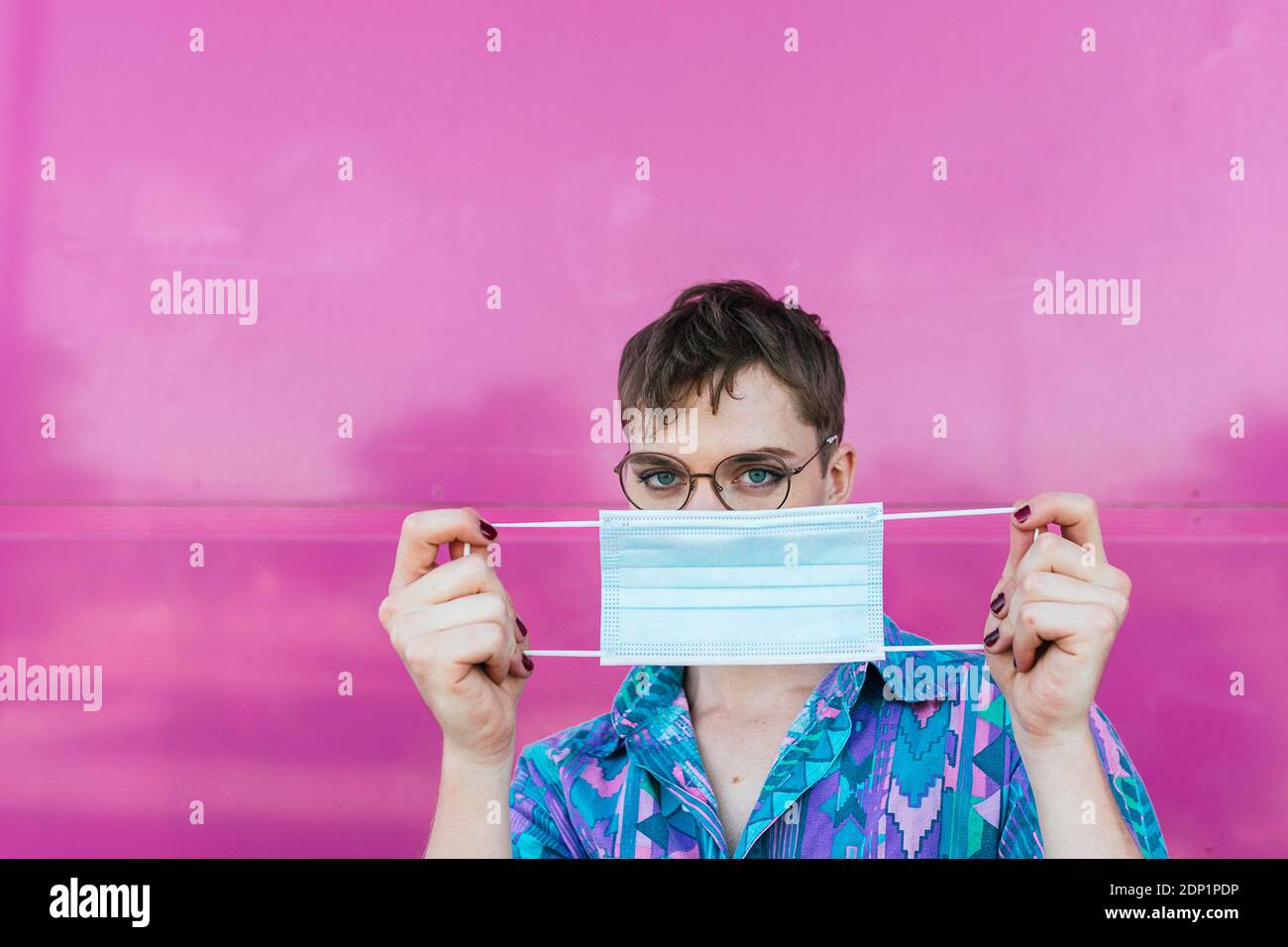 Portrait of non-binary person with face mask and pink glass wall ...