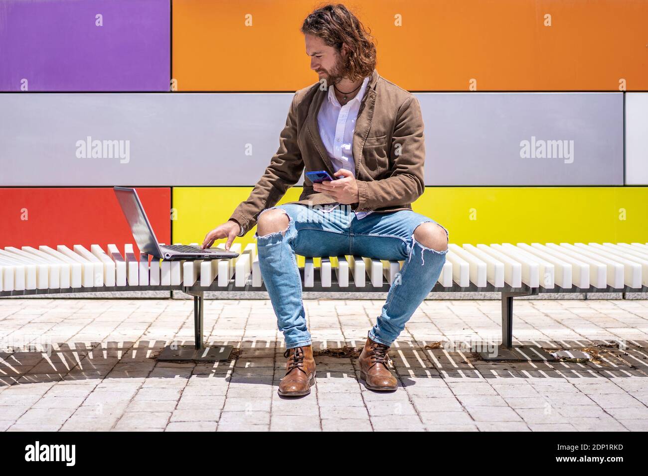 Young male professional holding smart phone while using laptop on bench against colorful wall Stock Photo