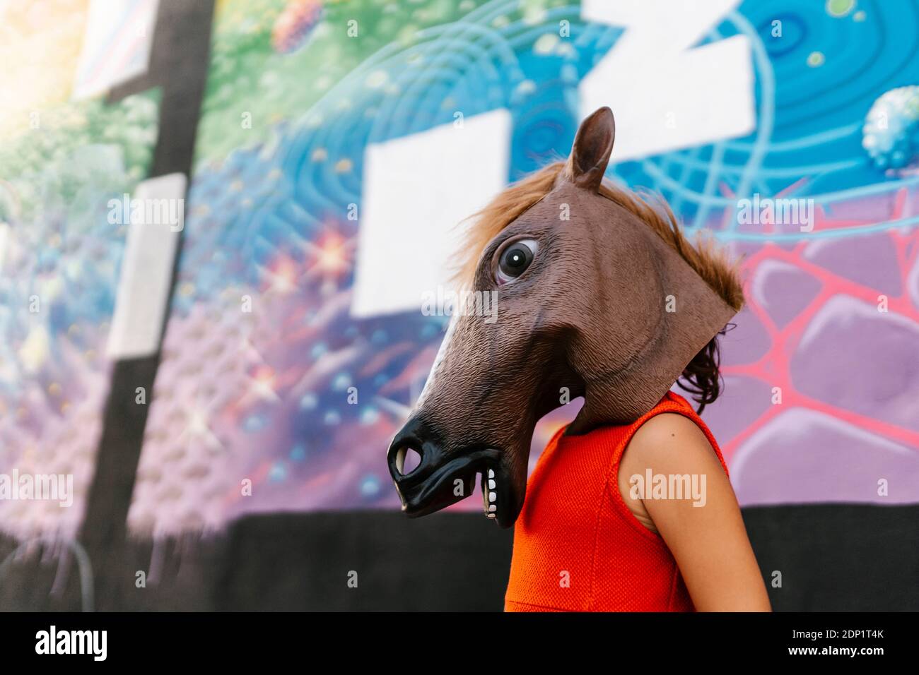 Little girl with a horse's head and a red dress next to a graffiti wall Stock Photo
