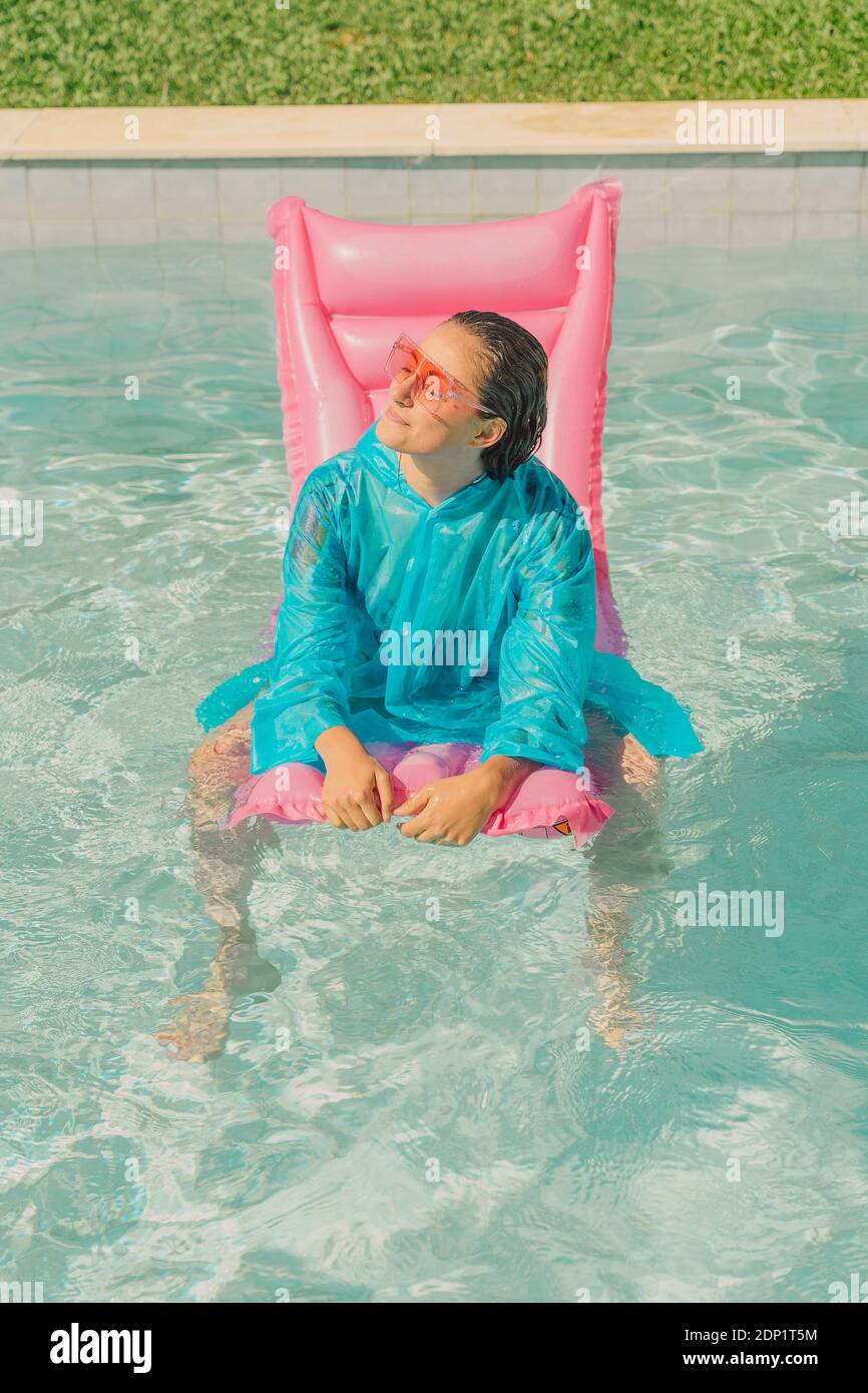 Woman wearing blue rain coat relaxing on pink airbed in swimming pool Stock Photo