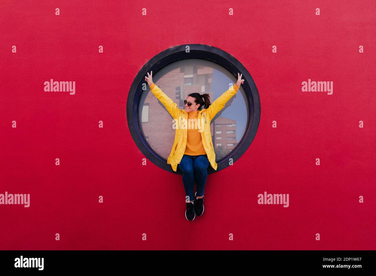 Woman in yellow raincoat sitting in porthole in red wall making victory sign Stock Photo