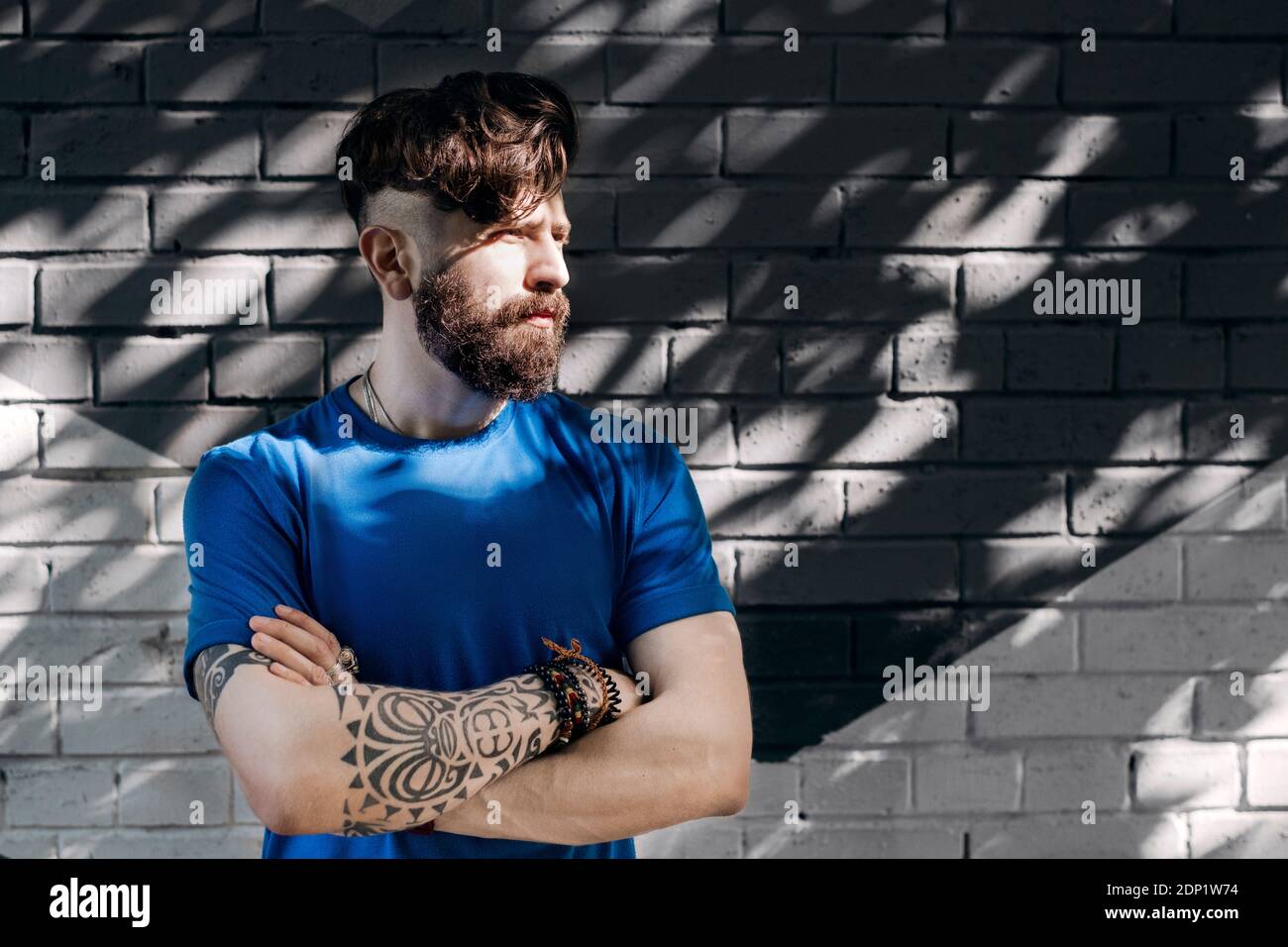 Young man with tattoo standing in front of brick wall with arms crossed Stock Photo