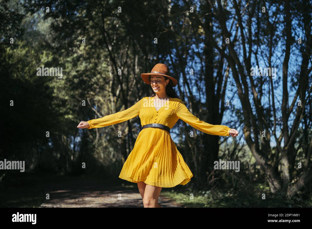 Happy young curly haired woman wearing brown hat and yellow dress walking with open arms and closed eyes Stock Photo