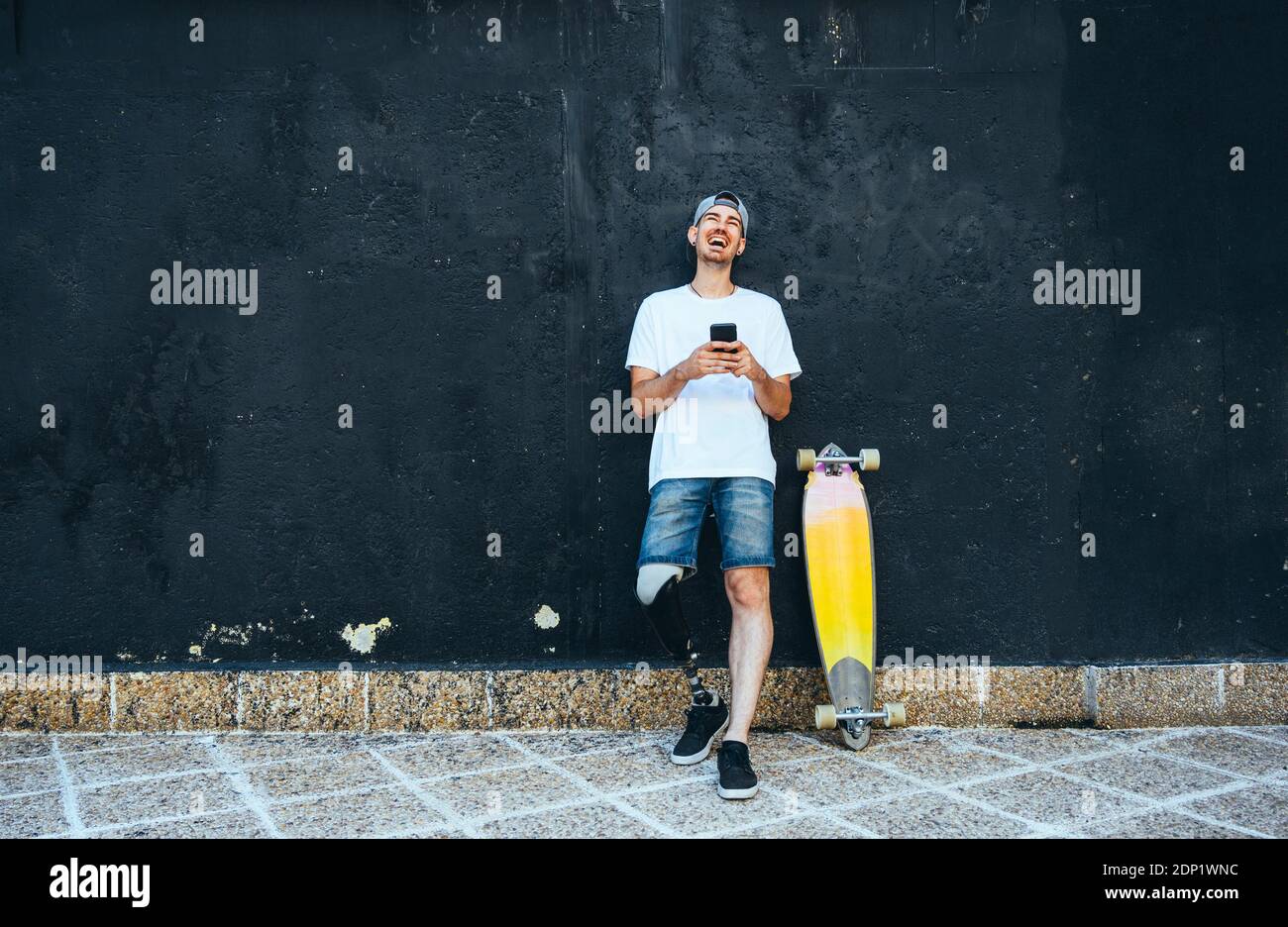 Laughing young man with leg prosthesis and skateboard using smartphone Stock Photo