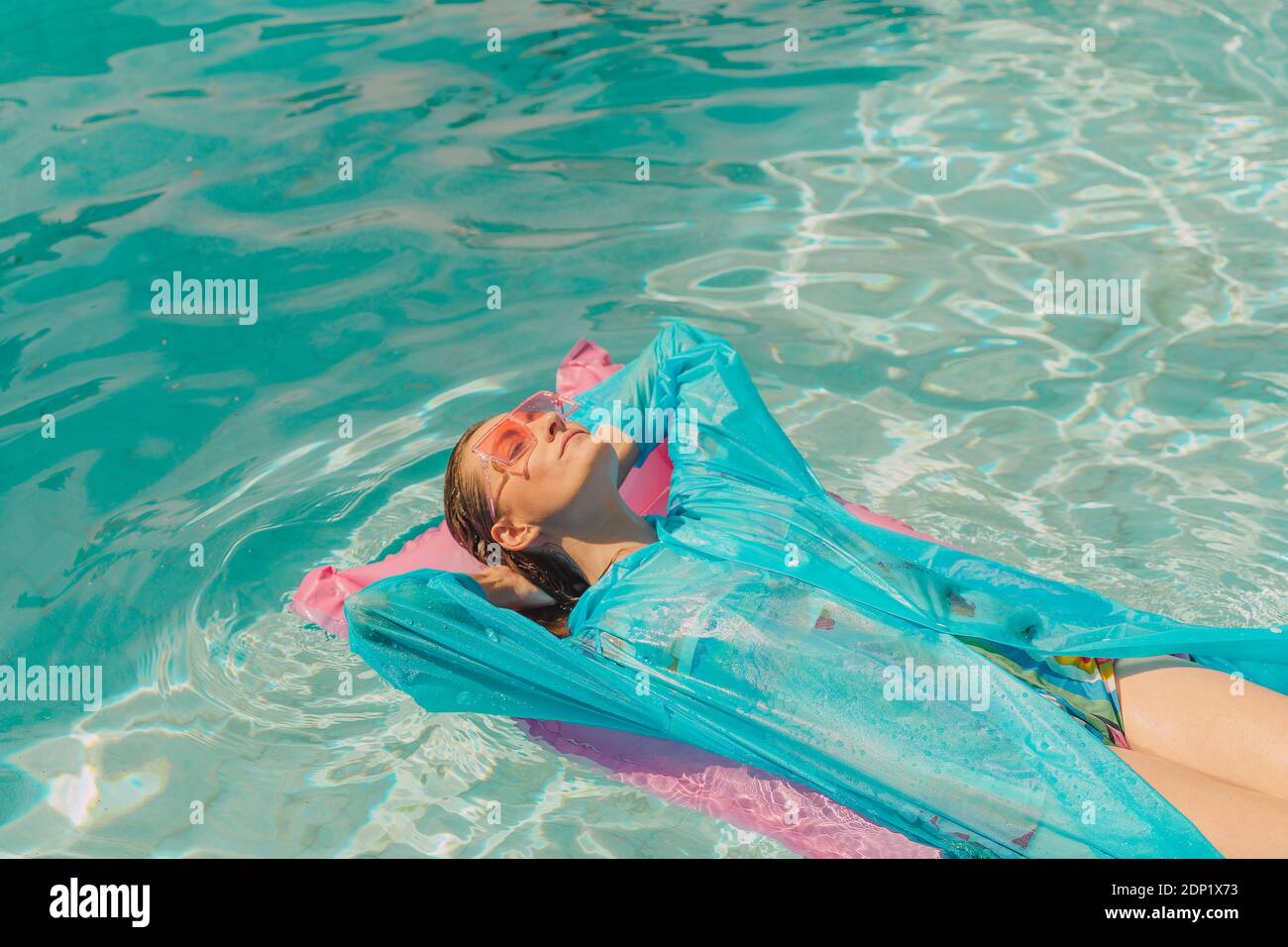 Woman wearing blue rain coat relaxing on pink airbed in swimming pool Stock Photo