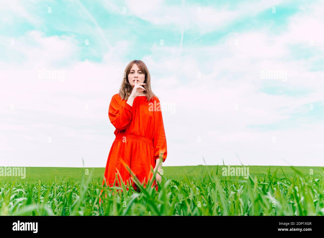 Portrait of woman wearing red dress standing in a field with finger on mouth Stock Photo