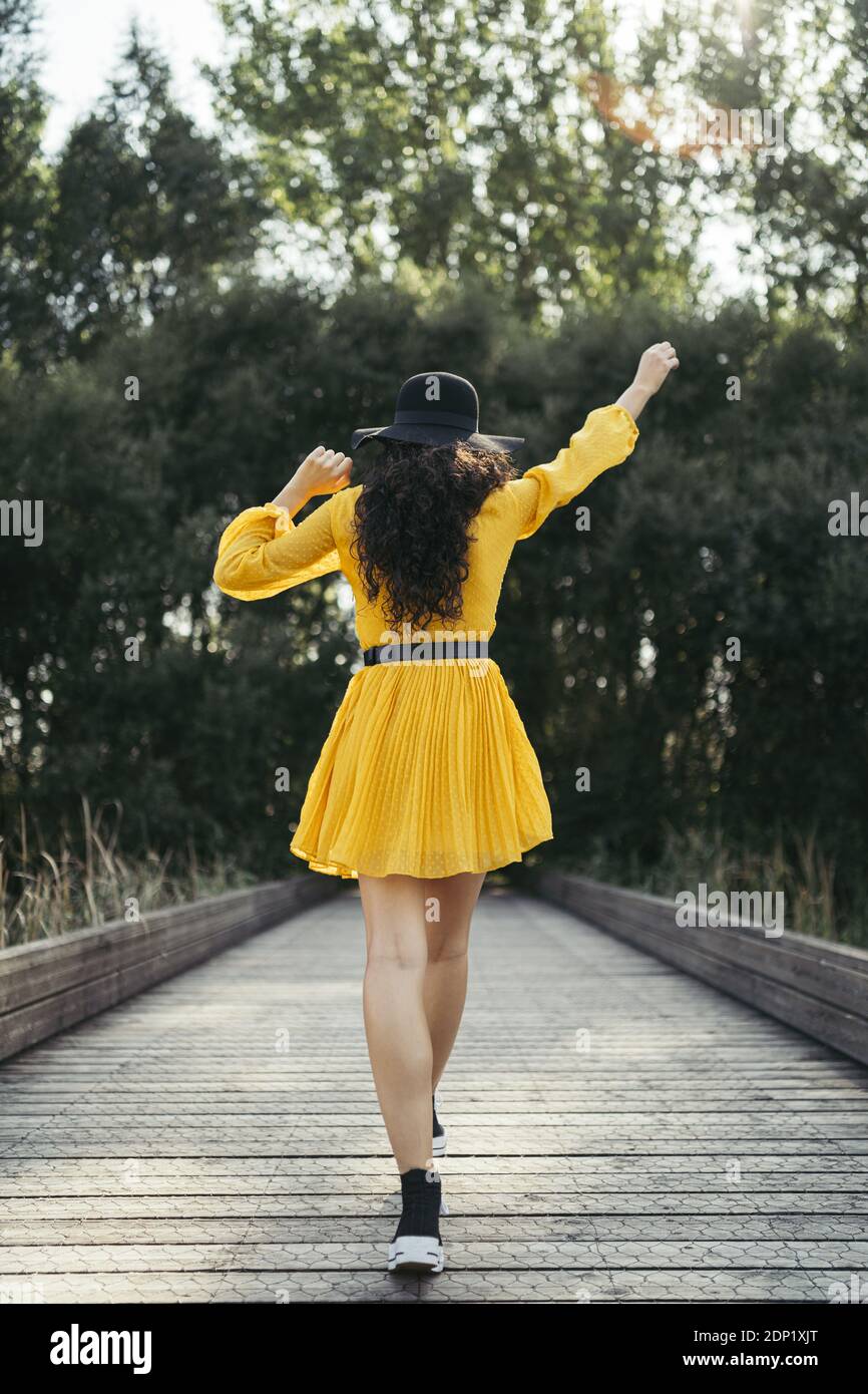 Rear view of a young woman wearing black hat and yellow dress enjoying while is walking along wooden bridge Stock Photo