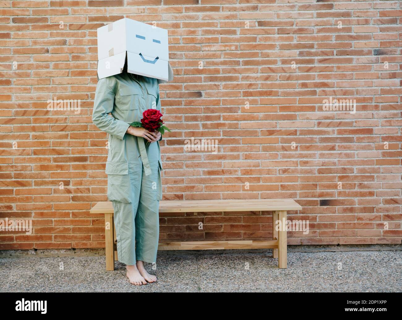Woman holding red rose, wearing cardboard box with happy face, standing by bench in front of brick wall Stock Photo