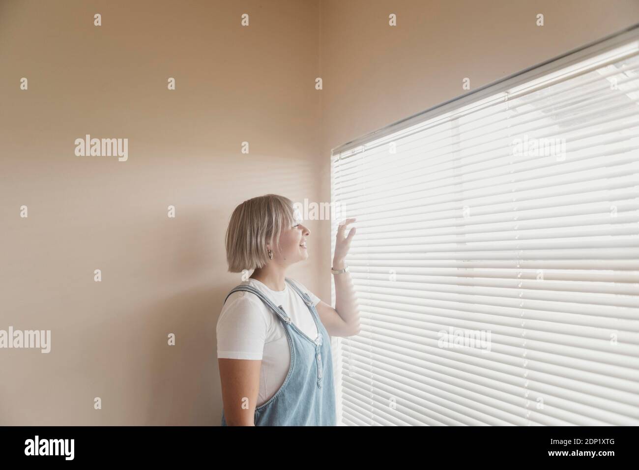 Happy young woman looking through blinds at the window Stock Photo
