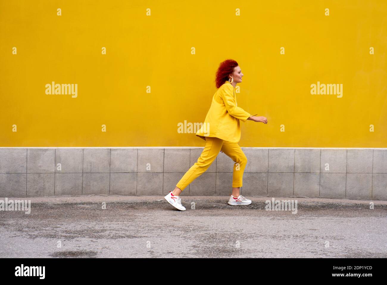 Energetic young woman in yellow suit running and jumping in front of yellow wall Stock Photo