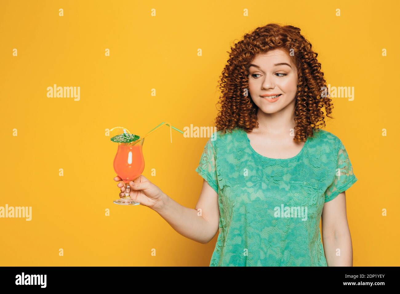 Curly red-haired woman in green t-shirt with orange tropical cocktail in hand over yellow background Stock Photo