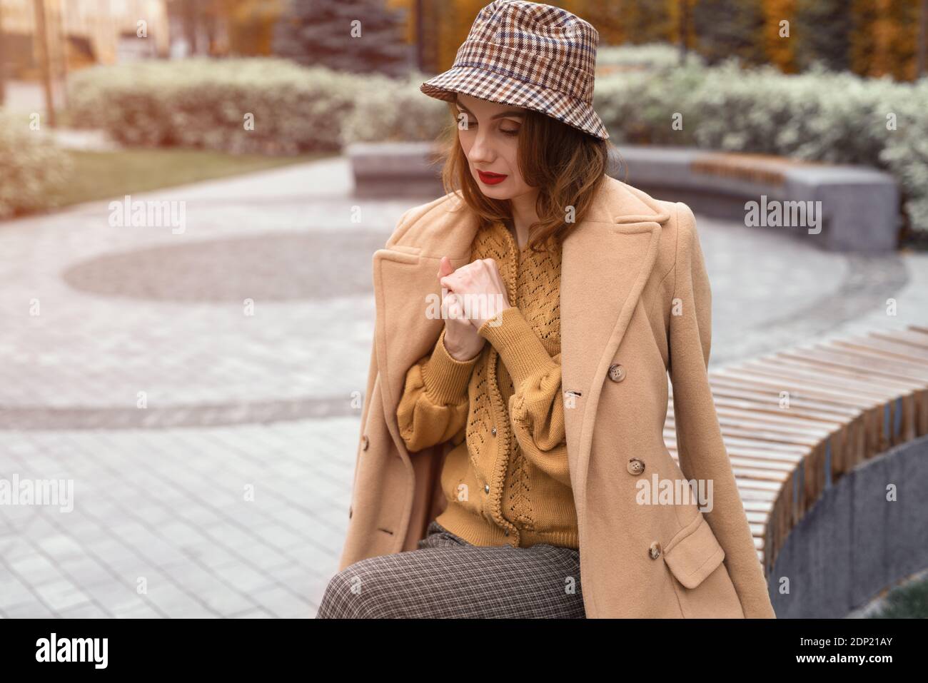 Parisian girl in beige coat and plaid panama hat siting on rounded bench waiting for her date or girlfriends. Autumn walk concept. Retro tinted photo Stock Photo