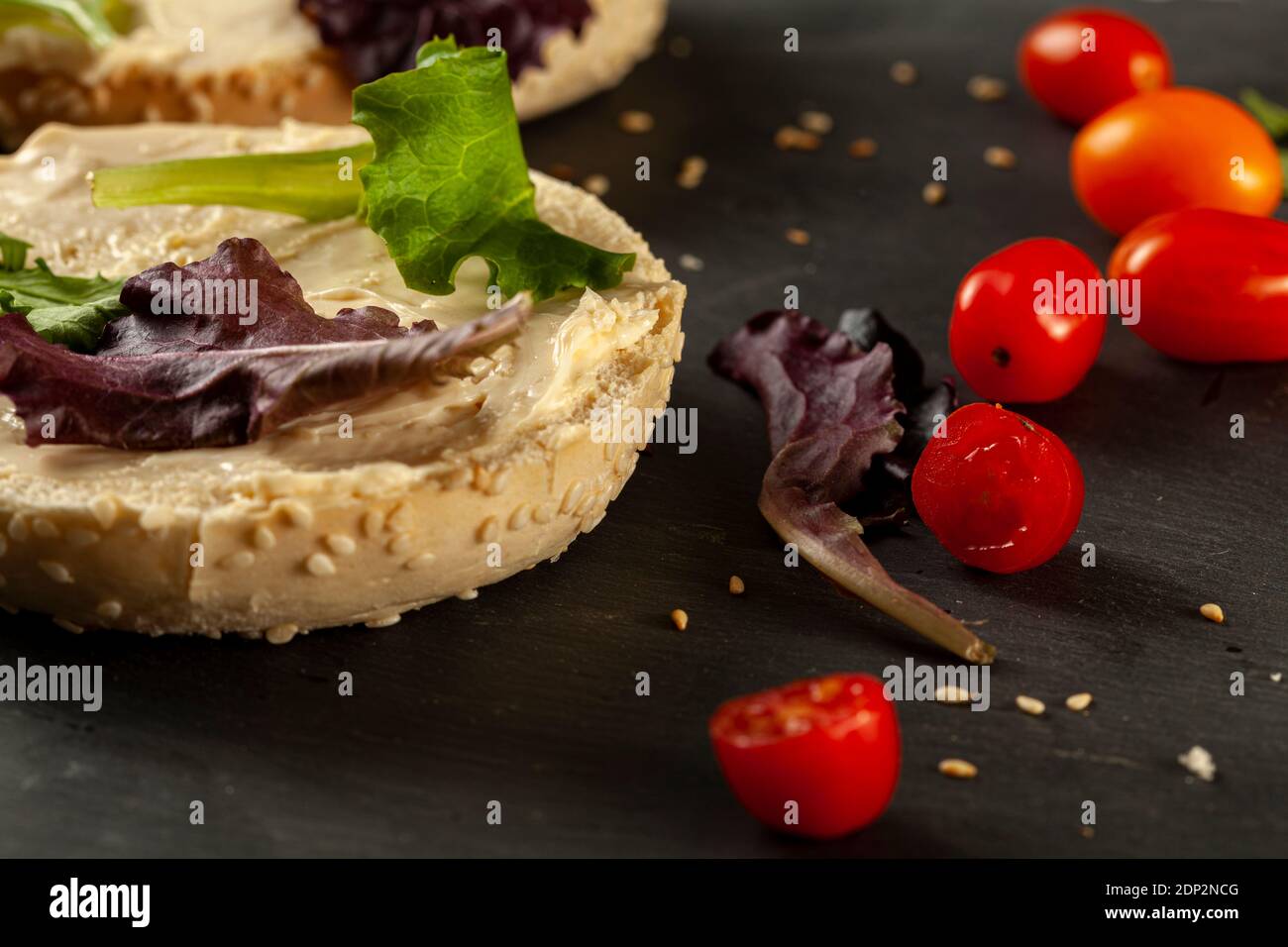 Toasted and sliced sesame bagel with creamy spread on it served with fresh greens (arugula) and cherry tomatoes on black wooden background. A healthy Stock Photo