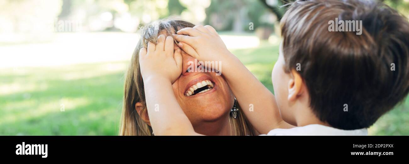 Woman playing with her child. Little boy covering the eyes of his mom. Mischiefs of al small kid getting fun in the park. Motherhood concept. Stock Photo