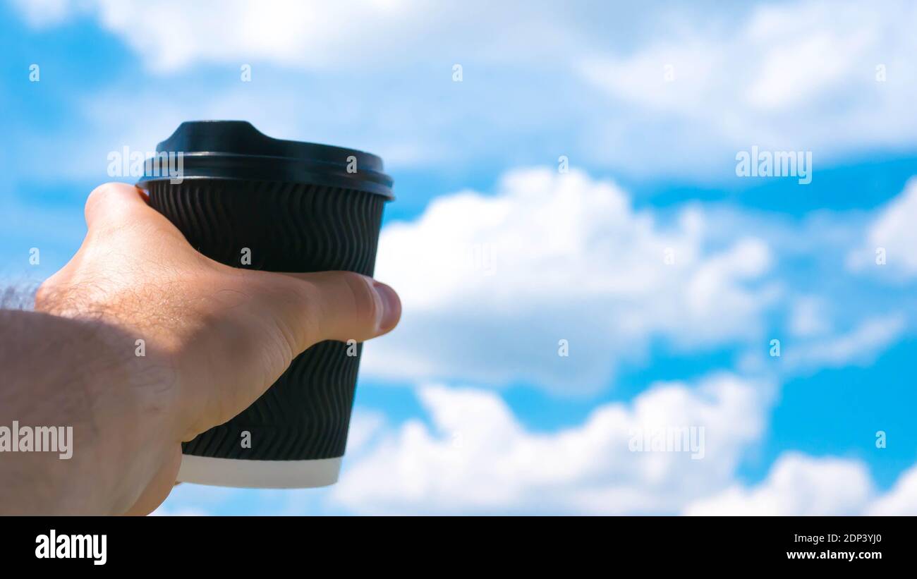 A glass of cappuccino,black coffee in a mans hand,the background of the sky is beautiful clouds and fluffy,close-up. Stock Photo