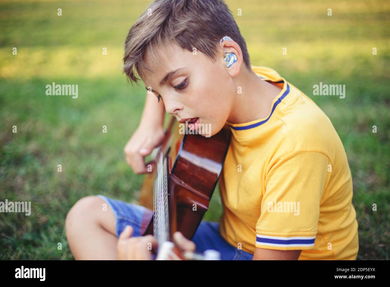 Hard of hearing preteen boy playing guitar and singing. Child with hearing aids in ears playing music and singing song in park. Hobby art activity for Stock Photo
