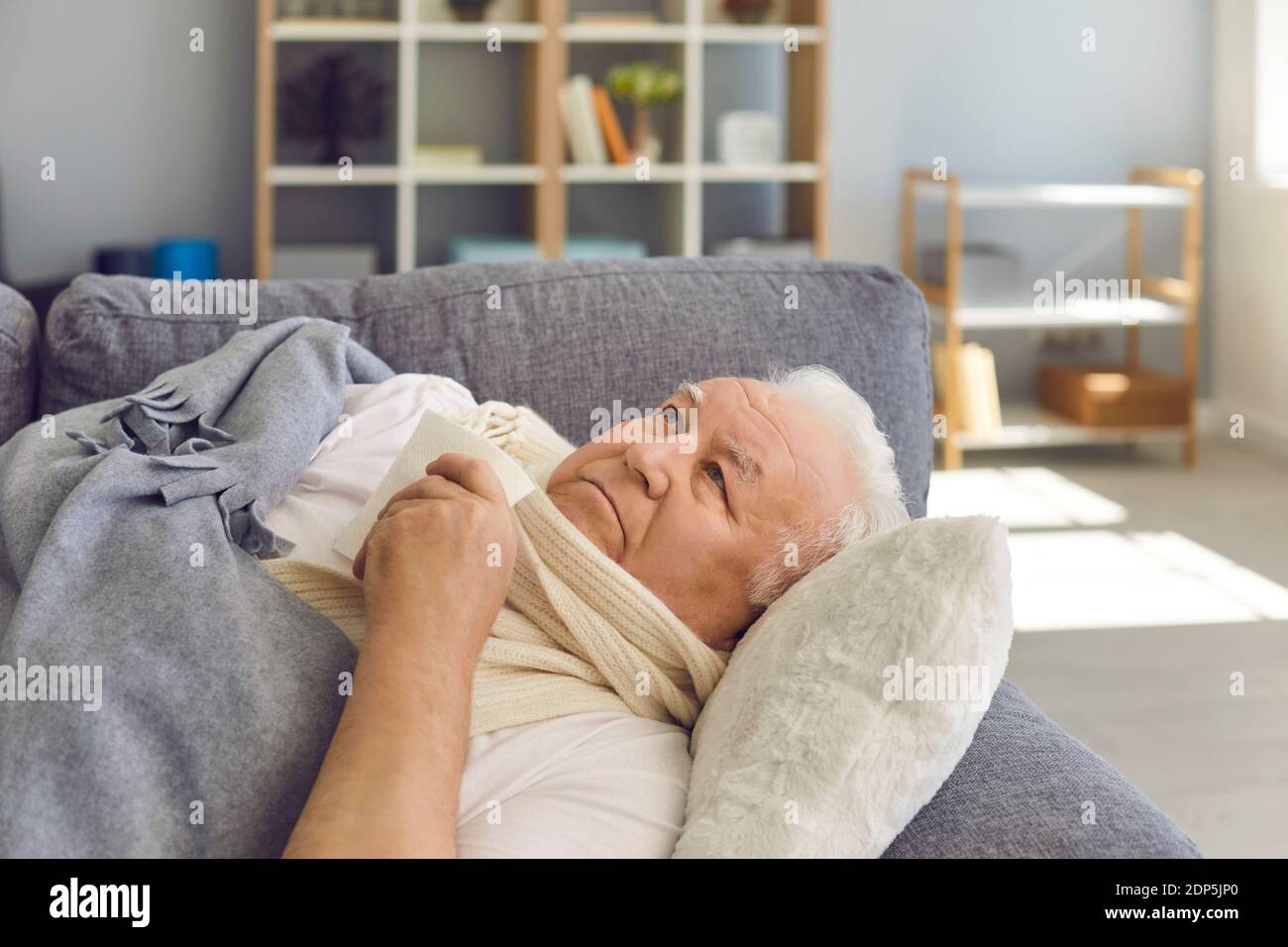 Sad senior man in scarf lying in bed with handkerchief under blanket and feeling ill Stock Photo